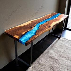 May include: A handcrafted wooden console table with a striking blue resin river design. The table features a live edge, showcasing the natural wood grain, and is supported by black metal legs. The table measures approximately 60 inches long.