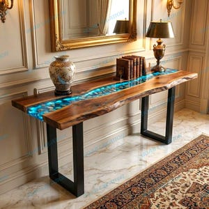 Puede incluir: Una consola de madera artesanal con un diseño de borde vivo. La mesa presenta un río de resina iluminada en azul incrustada con piedras. La mesa tiene patas de metal negro y está coronada por un jarrón decorativo y libros. La mesa mide aproximadamente 150 cm de largo.
