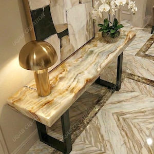 Può includere: Una moderna consolle con piano in pietra naturale nei toni crema, beige e oro, sostenuta da gambe in metallo nero. Una lampada dorata a forma di fungo è appoggiata sul tavolo. Un dipinto e un'orchidea in vaso sono sullo sfondo.