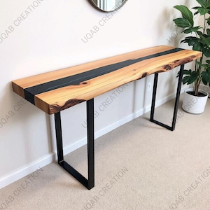 May include: A wooden console table with a natural wood grain finish and a black epoxy resin inlay. The table has black metal legs and is displayed against a white wall. The table is approximately 72 inches long.