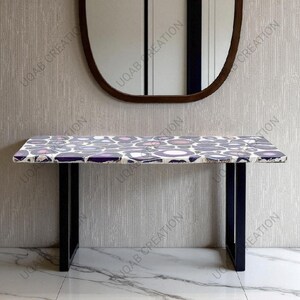 May include: A rectangular console table with a patterned top featuring purple, pink, and white circular designs. The table has black metal legs and is positioned in front of a wall with a mirror above it. The text "UQAB CREATION" is visible.