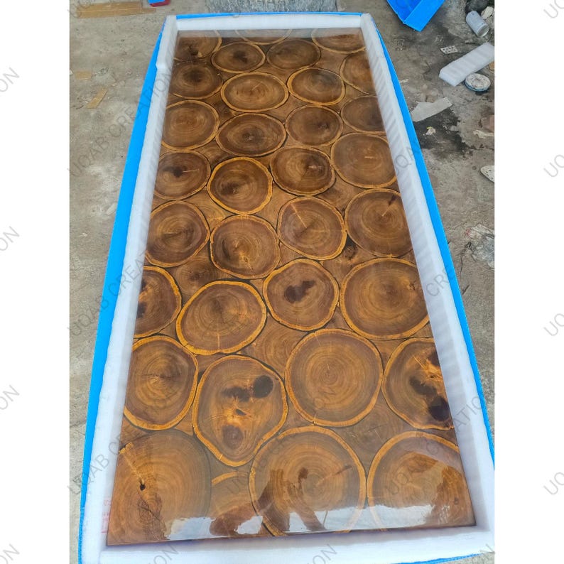 Puede incluir: Una encimera de mesa rectangular de madera con acabado de resina transparente. La encimera est&aacute; hecha de numerosas rodajas de madera circulares, cada una de ellas con patrones de vetas &uacute;nicos y tonos marrones c&aacute;lidos. La mesa est&aacute; encerrada en un marco protector azul y blanco.