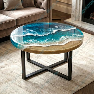 Op de afbeelding: Ronde salontafel met een hars oceaan ontwerp. Het tafelblad heeft een realistische weergave van oceaangolven in tinten blauw en wit, met een zandstrandrand. De tafel heeft een zwart metalen onderstel.