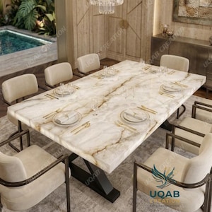 Puede incluir: Una mesa de comedor rectangular con tablero de mármol veteado en blanco y dorado sobre una base de metal negro, rodeada de ocho sillas tapizadas en color crema con marcos de metal. Cada cubierto incluye un plato blanco, cubiertos y un vaso.