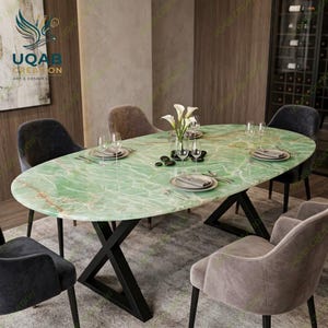Puede incluir: Una mesa de comedor ovalada con una superficie de mármol verde pulido con vetas blancas y una base de metal negro en forma de X. La mesa está dispuesta con platos blancos, cristalería transparente y cubiertos, rodeada por cuatro sillas tapizadas.