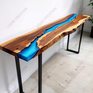May include: A long, rectangular wooden console table with a river of bright blue resin running down the center. The wood has a natural edge and a rich brown color. The table is supported by black metal legs.