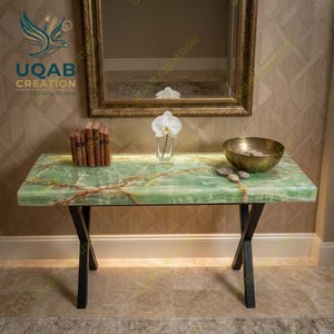 Peut inclure: Une console rectangulaire avec un plateau en onyx vert clair poli aux veines brunes, reposant sur une base en métal noir en forme de X. Sur la table se trouvent six livres verticaux, un vase en verre transparent avec une orchidée blanche et un bol doré.