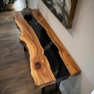 Op de afbeelding: Een handgemaakte houten console met een live edge design. De tafel heeft een donkere, glanzende zwarte epoxyhars rivier die door het midden loopt, contrasterend met de natuurlijke houtnerf. Hij wordt ondersteund door zwarte metalen poten.