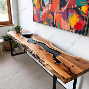 Puede incluir: Una consola de madera de borde vivo con un río de resina epoxi negra en el centro. La mesa está soportada por patas de metal negro. Un cuenco blanco se encuentra encima. La mesa mide aproximadamente 183 cm de largo.