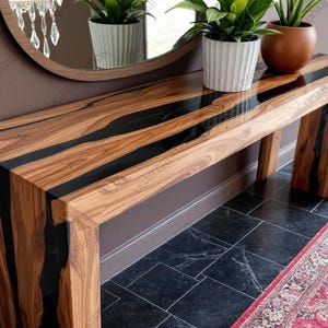 May include: A wooden console table with a natural wood grain finish and black resin accents. The table has a rectangular top and four sturdy legs. The wood is a warm brown color, and the resin is a glossy black. The table is a modern piece of furniture.