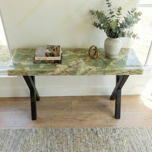 Puede incluir: Una mesa de centro rectangular de ónix verde con patas de metal negro. La mesa tiene una superficie pulida con vetas naturales. Una pila de libros, un objeto decorativo dorado y un jarrón con vegetación están sobre la mesa.