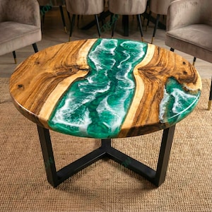 Può includere: Tavolino rotondo con piano in legno e un fiume di resina verde e bianca. Le sezioni in legno sono di colore marrone chiaro. Il tavolo ha una base in metallo nero.