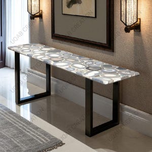 May include: A long, rectangular console table with a patterned agate stone top in shades of gray, white, and blue. The table has a black metal frame and is placed against a neutral-toned wall. Decorative wall sconces flank a framed artwork.