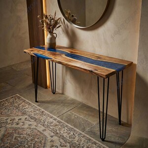 Op de afbeelding: Een houten console tafel met een live edge design en een blauwe hars rivier. De tafel heeft zwarte hairpin poten. Een vaas met gedroogde bloemen staat op de tafel. De tafel is ongeveer 152 cm lang.