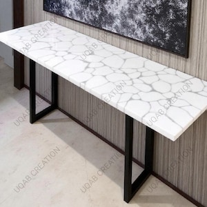 May include: A long, rectangular console table with a white, patterned top and black metal legs. The white surface features a gray, organic pattern. The table is a modern design, suitable for entryways or living spaces.