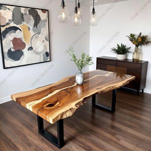May include: A wooden dining table with a live edge design, showcasing natural wood grain. The table has a rectangular shape and a black metal base. The table is in a dining room setting with a painting and a cabinet.