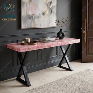 May include: A rectangular console table with a polished pink quartz top and a black metal X-frame base. On the table sits a round tray with a glass candle holder, a stack of two books, and a black ceramic vase containing dried branches.
