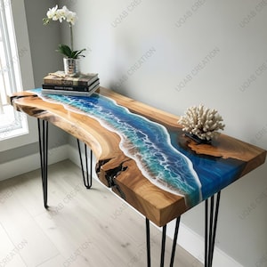 Puede incluir: Una consola de madera con un diseño de océano de resina. La mesa presenta un borde de madera natural y patas de horquilla negras. El diseño de resina incorpora tonos de azul, blanco y beige, que se asemejan a una escena de playa. Un coral decorativo y libros están sobre la mesa.