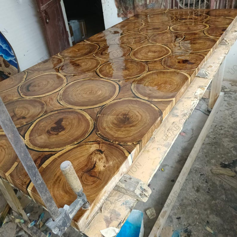 Puede incluir: Una mesa de madera rectangular grande con un acabado de resina transparente y brillante. La superficie de la mesa est&aacute; hecha de numerosas rodajas de madera circulares, que muestran la veta natural y las variaciones de color de la madera. La mesa est&aacute; soportada por un marco de madera.