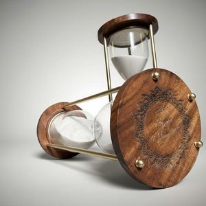 Op de afbeelding: Een zandloper met een houten frame en decoratieve ronde uiteinden. De glazen kamers bevatten wit zand, ondersteund door messingkleurige metalen staven. De houten uiteinden zijn gegraveerd met een gedetailleerd patroon.