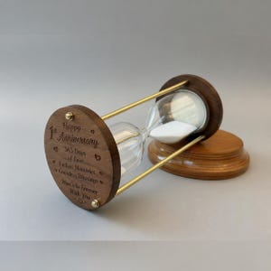 Pode incluir: Uma ampulheta de madeira com uma câmara de vidro transparente preenchida com areia branca. A ampulheta tem uma estrutura e base de madeira castanha escura, com suportes de metal dourado. O texto "Happy 1st Anniversary" está gravado na base de madeira.