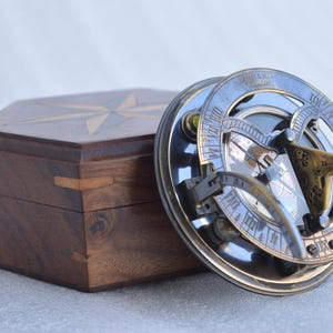 May include: A vintage sundial with a compass and a wooden box. The sundial is made of metal with a brass finish and features intricate details. The wooden box is dark brown with a star design on the lid.