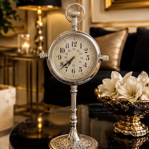 Peut inclure: Une horloge de table de style vintage, de couleur argentée, avec un cadran rond et des chiffres romains. L'horloge a une base décorative et une boucle en haut. Elle est posée sur une table noire avec un bord doré. Un bol doré avec des fleurs blanches est à proximité.