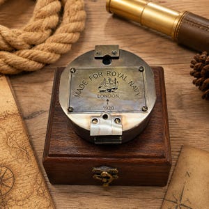 Peut inclure: Une boussole en laiton vintage, marquée "Made for Royal Navy London 1920", est posée sur une boîte en bois avec un fermoir en laiton. Un thème nautique est mis en place avec une corde, une longue-vue et une carte en arrière-plan. La boussole a une gravure détaillée d'un navire.