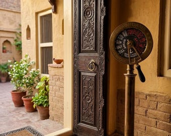 May include: An open, ornately carved dark wooden door with a brass handle. A vintage brass telegraph is positioned to the right. The doorway leads to a courtyard with terracotta tiles. Potted plants and a patterned rug are visible.