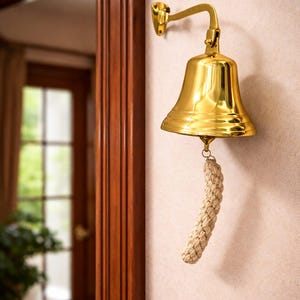 May include: A polished brass bell fixed to a wall. It features a curved arm and a rope clapper. The bell is likely used as a decorative item or a signaling device. The bell's dimensions are approximately 15 cm in height.