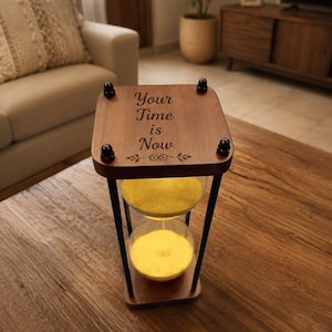 Könnte beinhalten: Eine hölzerne Sanduhr mit einem dunkelbraunen Rahmen und schwarzen Akzenten. Auf der Oberseite der Sanduhr steht der Text "Your Time is Now". Der Sand im Inneren ist gelb. Die Sanduhr steht auf einem Holztisch.
