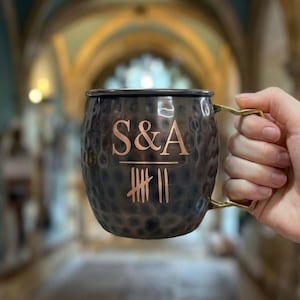 May include: A dark, hammered metal mug with a metallic gold-toned handle. The front of the mug features the engraved letters S&A above a horizontal line and a tally mark design consisting of five vertical lines with a diagonal strike and two extra lines.