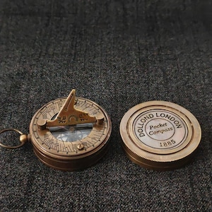 May include: A vintage brass pocket compass with a sundial and a separate lid. The compass is open, revealing the dial and needle. The lid is closed and has the text "DOLLOND LONDON Pocket Compass 1885". The compass is on a dark gray textured surface.