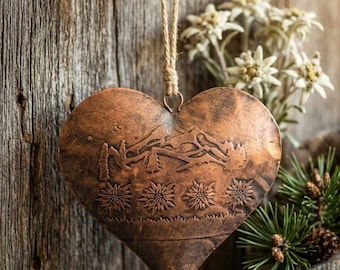 Könnte beinhalten: Ein herzförmiges Kupferornament mit einer geprägten Bergkette und einer Reihe von vier Edelweißblüten. Das Metallstück weist eine verwitterte, dunkle Patina auf und hängt an einer Schlaufe aus natürlichem Jutegarn an einem kleinen Metallring.