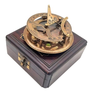 May include: A vintage brass sundial with Roman numerals and a gnomon, resting on a dark wooden box. The sundial features intricate details and a small green gemstone. The box has a brass clasp.