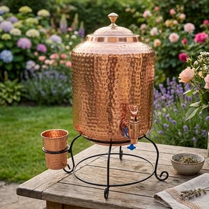 May include: A hammered copper water dispenser with a matching cup, featuring a lid with a knob handle and a blue spigot. The dispenser rests on a black metal stand with curved legs, designed to hold the vessel and the cup securely in place.