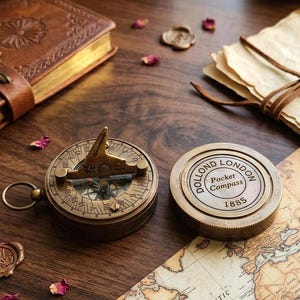 Pode incluir: Bússola de bolso e bússola de sol em latão antigo com um diário encadernado em couro e um mapa vintage. As bússolas são marcadas "DOLLOND LONDON Pocket Compass 1885". Pétalas de rosa espalhadas adicionam cor.