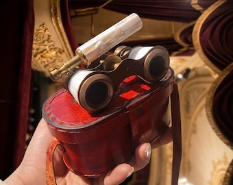 May include: Antique opera glasses featuring a brass frame with white mother-of-pearl inlays and an extendable handle. The binoculars rest on a red leather carrying case with a matching leather strap, designed for viewing performances.