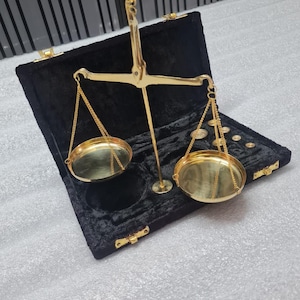 May include: A gold-coloured balance scale with two circular pans suspended by chains. The scale is inside a black velvet-lined case with gold-coloured hardware. The scale is designed for weighing small objects.