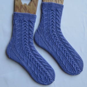 Könnte beinhalten: Ein Paar periwinkelfarbene Strickstrümpfe mit Zopfmuster. Die Socken werden auf Holzformen präsentiert. Das Zopfmuster verläuft von der Spitze bis zum Bündchen der Socken. Die Socken sind aus weichem, strukturiertem Garn gefertigt.