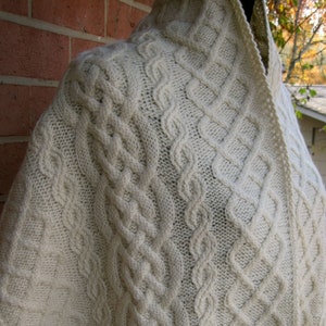 Pode incluir: Um xale de malha branca com um padrão de malha de cabo. O xale está drapeado sobre o ombro de um manequim.