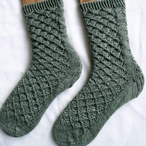 May include: A pair of sage green knit socks featuring a textured diamond lattice pattern across the leg and foot, with a ribbed cuff at the top and a smooth, plain knit finish on the toe and heel sections, crafted from a heathered yarn.