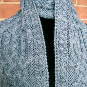 May include: A light blue knitted scarf with a cable knit pattern. The scarf is folded in half and draped over a black surface.