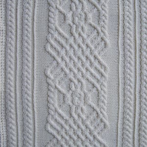 May include: Close-up of a white knitted fabric with a cable knit pattern. The fabric has a textured surface and a soft, cozy look.