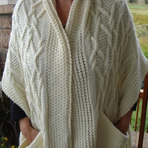 May include: A white knitted shawl with a cable knit pattern and two pockets.