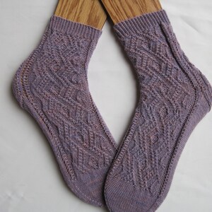 May include: A pair of purple and pink knitted socks with a cable knit pattern. The socks are displayed on wooden sock blockers.