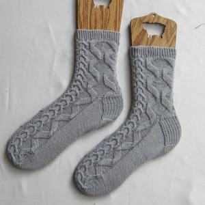 May include: A pair of gray knitted socks with a cable knit pattern. The socks are displayed on wooden sock blockers.