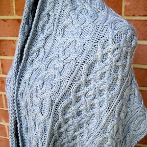 May include: A light blue knitted shawl with a cable knit design. The shawl is draped over a shoulder, showcasing the intricate pattern and texture of the yarn. The background is a red brick wall.