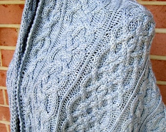 May include: A pale blue knitted shawl featuring a cable knit pattern. The shawl is draped over a shoulder, highlighting the detailed texture of the yarn. The background is a red brick wall.