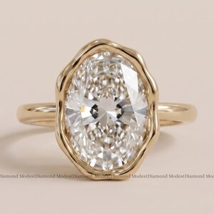 May include: A gold ring featuring a large, oval-cut clear gemstone set in a bezel with a wavy, organic-shaped edge. The band is smooth and polished gold, providing a clean contrast to the textured, irregular frame surrounding the central stone.